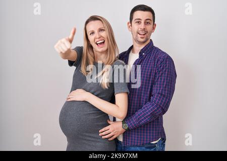 Jeune couple s'attendant à un bébé debout sur fond blanc approuvant faire un geste positif avec la main, pouces vers le haut souriant et heureux pour le succès. winn Banque D'Images