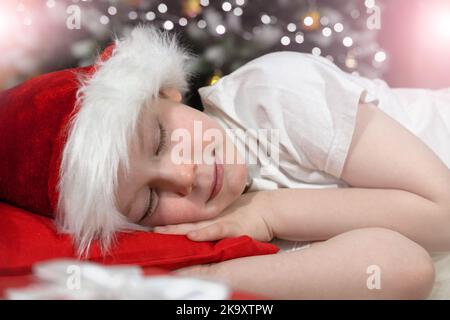 Rêves de Noël d'un enfant. Un enfant dans un chapeau de Père Noël dort dans le lit près d'un arbre de Noël avec des lumières de Noël, en attendant le cadeau du Père Noël. Banque D'Images