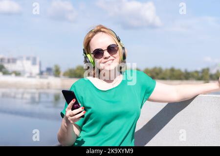 Une femme souriante avec un casque tenant un téléphone dans la main écoute de la musique sur la rive. Une jeune fille blogueuse portant un casque à l'aide d'un smar Banque D'Images