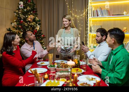 Divers amis en visite pour Noël célébrant les vacances du nouvel an dîner et se réjouir de s'amuser, groupe de personnes mangeant et buvant du vin à la table à la maison près de l'arbre de Noël. Banque D'Images