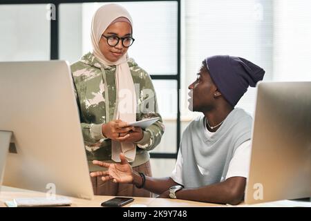 Deux jeunes programmeurs multiculturels en vêtements décontractés ayant une discussion sur de nouveaux points de projet tandis que la femme musulmane écoute un collègue masculin Banque D'Images