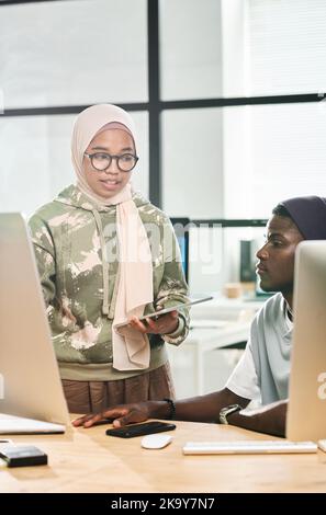 Une jeune femme d'affaires musulmane dans le hijab expliquant quelque chose à un collègue afro-américain assis entre deux moniteurs Banque D'Images