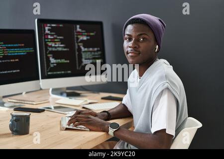 Jeune responsable de l'assistance informatique afro-américain confiant regardant la caméra tout en étant assis sur le lieu de travail devant les moniteurs d'ordinateur Banque D'Images