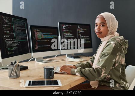Jeune spécialiste informatique moderne en hijab et sweatshirt, qui décode les informations sur les écrans d'ordinateur tout en étant assis devant trois moniteurs Banque D'Images