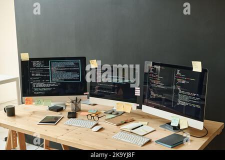 Trois écrans d'ordinateur avec données codées et feuilles de notes adhésives sur leurs écrans sur le lieu de travail de l'ingénieur informatique moderne dans le bureau Banque D'Images