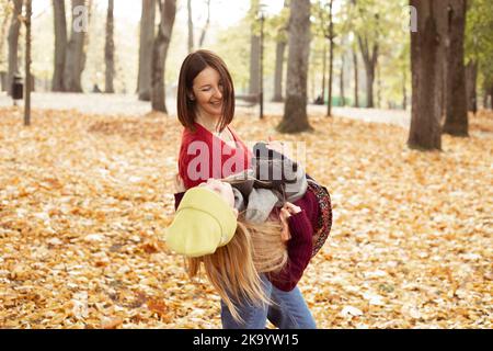 Une jeune femme joyeuse tient dans les mains mentant une fille dans un chapeau et un foulard. Bonne mère et fille dans les sweaters tricotés confortables ont le plaisir et jouer en automne doré Banque D'Images