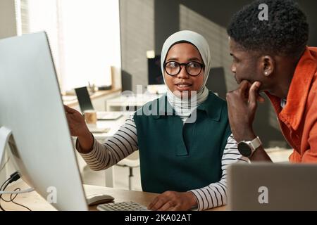 Jeune femme musulmane confiante dans le hijab pointant vers l'écran d'ordinateur et regardant un collègue mâle afro-américain pendant la présentation Banque D'Images