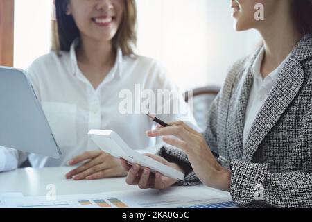 Deux jeunes femmes d'affaires asiatiques utilisant leurs tablettes informatiques avec des calculatrices et des documents pour planifier des stratégies de marketing et des investissements à tirer profit Banque D'Images