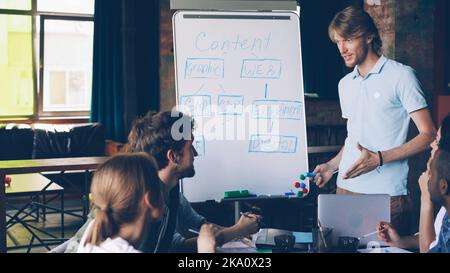Beau jeune homme fait une présentation créative pour l'équipe multiraciale en utilisant le tableau blanc, il pointe sur le texte avec un marqueur et parle. Concept de formateur commercial et de bureau. Banque D'Images