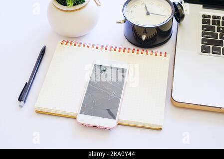 Vue de dessus d'une table de bureau blanche avec ordinateur portable, réveil noir et smartphone craquer sur ordinateur portable Banque D'Images