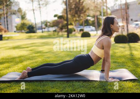Vue latérale d'une femme flexible qui s'étire la colonne vertébrale, le dos, sur le tapis, en commençant le matin par des exercices de yoga dans le parc de la ville. Femme de fitness. Soleil du matin Banque D'Images