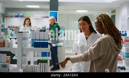 Pharmacien asiatique parlant à divers clients atteints de maladie, en donnant des produits pharmaceutiques à partir des étagères de pharmacie. Employé donnant de l'aide médicale aux gens, expliquant les médicaments dans la pharmacie. Banque D'Images