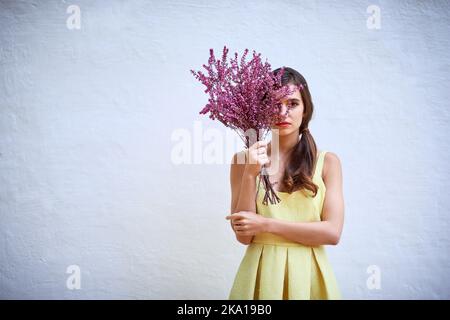 Où les fleurs fleurissent ainsi sa beauté. Portrait studio d'une jeune femme confiante se cachant derrière un bouquet de fleurs tout en se tenant contre un gris Banque D'Images