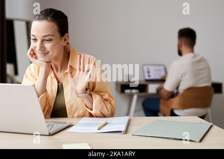Un jeune étudiant souriant se fait la main devant un tuteur lors d'une leçon en ligne ou d'un séminaire en ligne tout en étant assis devant l'écran d'un ordinateur portable à la maison Banque D'Images