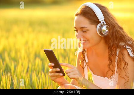 Une femme heureuse dans un champ qui écoute de la musique avec le téléphone au coucher du soleil Banque D'Images