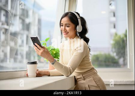 Jolie jeune femme asiatique charmante dans des vêtements décontractés près de la fenêtre, en écoutant de la musique par le biais d'écouteurs, en choisissant une liste de lecture de musique sur elle Banque D'Images