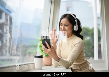 Belle jeune femme asiatique vêtue de vêtements décontractés, portant un casque, se balader, parlant en vidéo sur son smartphone. Banque D'Images