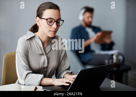 Jeune femme brune sérieuse en vêtements décontractés et lunettes de vue assis devant un ordinateur portable et en tapant pendant l'exercice Banque D'Images