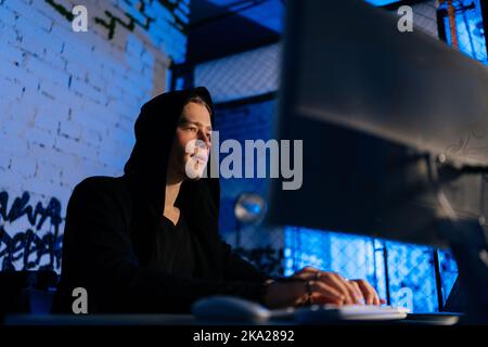 Vue rapprochée à faible angle de l'homme pirate qui vole des informations, se préparant à briser le mot de passe assis au bureau dans la pièce sombre avec des lumières néon bleues Banque D'Images