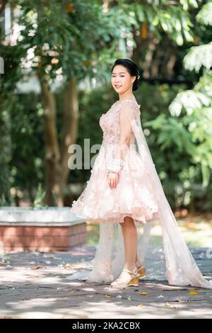 Fille asiatique dans une vieille rose rose couleur genou longueur robe molletonnée avec le talon élevé des chaussures à bride arrière et le fil de cérémonie sur son poignet. Elle est plus lumineuse Banque D'Images