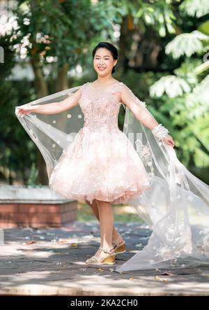 Fille asiatique dans une vieille rose rose couleur genou longueur robe molletonnée avec le talon élevé des chaussures à bride arrière et le fil de cérémonie sur son poignet. Elle est plus lumineuse Banque D'Images