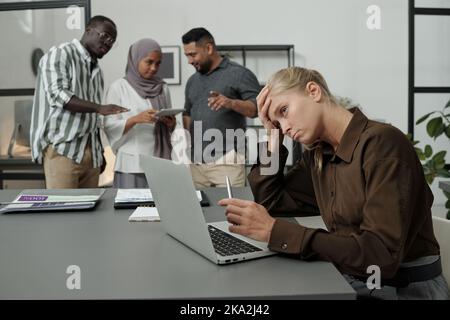 Jeune femme d'affaires fatiguée essayant de se concentrer pendant le travail sur le projet tandis que le groupe de collègues multiculturels l'intimide Banque D'Images