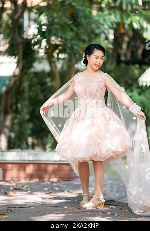 Fille asiatique dans une vieille rose rose couleur genou longueur robe molletonnée avec le talon élevé des chaussures à bride arrière et le fil de cérémonie sur son poignet. Elle est plus lumineuse Banque D'Images