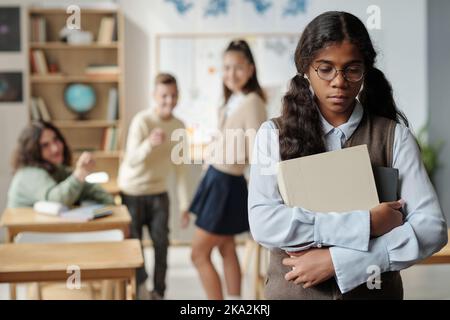 Avant l'adolescence bouleversé écolière avec des livres debout devant l'appareil photo avec l'expression offensé contre trois camarades de classe l'intimidant Banque D'Images