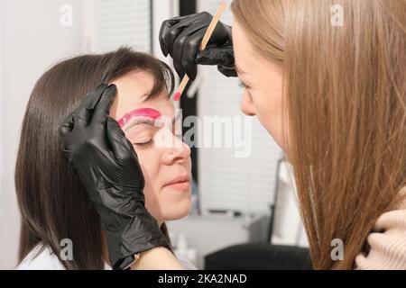 Jeune femme brunette ayant une procédure de correction des sourcils dans le salon de beauté. Jeune esthéticienne appliquant de la cire chaude sur le visage du client Banque D'Images