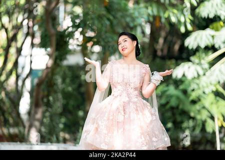 Fille asiatique dans une vieille rose rose couleur genou longueur robe molletonnée avec le talon élevé des chaussures à bride arrière et le fil de cérémonie sur son poignet. Elle est plus lumineuse Banque D'Images