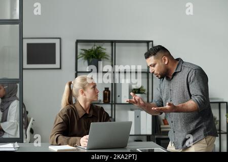 Jeune femme d'affaires blonde assise sur le lieu de travail devant un ordinateur portable et regardant un patron irrité expliquer ses points de travail Banque D'Images
