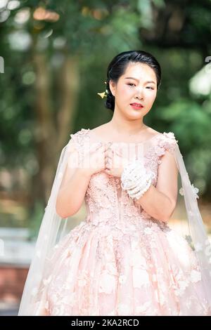 Fille asiatique dans une vieille rose rose couleur genou longueur robe molletonnée avec le talon élevé des chaussures à bride arrière et le fil de cérémonie sur son poignet. Elle est plus lumineuse Banque D'Images