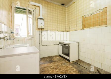 Vieille cuisine sale et délabrée avec un sol sombre, des carreaux blancs et du papier peint jaune sur les murs, une vieille cuisinière solitaire et un chauffe-gaz Banque D'Images