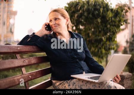 Bonne femme d'affaires mûre parlant sur un smartphone tenant un ordinateur portable Banque D'Images