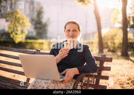 Bonne femme d'affaires senior tenant un ordinateur portable et de la crème glacée dans le parc Banque D'Images