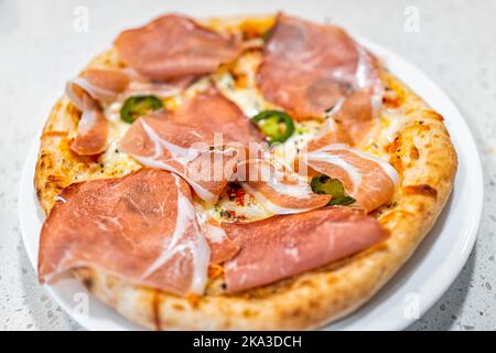 Macro-gros plan de jambon de porc cuit au four, fromage mozzarella fondu et poivrons jalapeno verts sur la tarte à la pizza sur la plaque blanche sur la table de cuisine Banque D'Images