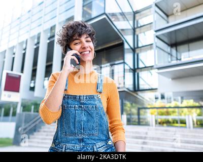 Femme ethnique aux cheveux bouclés regardant l'appareil photo heureusement en parlant sur smartphone près du bâtiment sur la rue de la ville Banque D'Images
