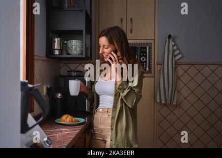 Bonne femme hispanique avec une tasse de café souriant et parlant sur le téléphone portable tout en se tenant près du comptoir avec croissant pendant le petit déjeuner le matin à ki Banque D'Images
