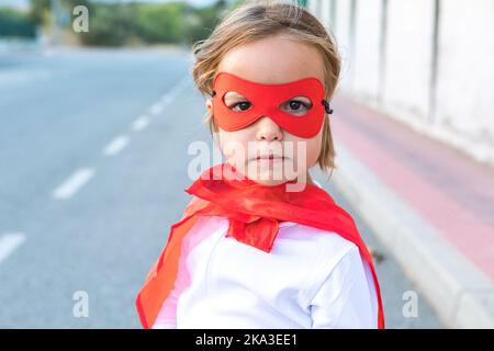 Garçon en costume de super-héros et masque regardant l'appareil-photo en se tenant sur la route asphaltée Banque D'Images