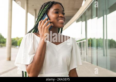 Content jeune femme afro-américaine avec des cheveux tressés verts parlant au-dessus de smartphone tout en étant debout dans la rue Banque D'Images