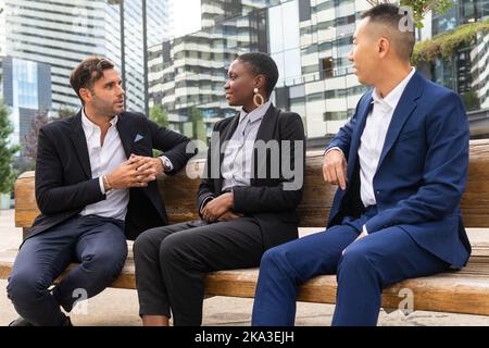 Collègues multiraciaux positifs en costume habillé assis sur un banc en bois près du centre d'affaires moderne et bavardant pendant la pause Banque D'Images