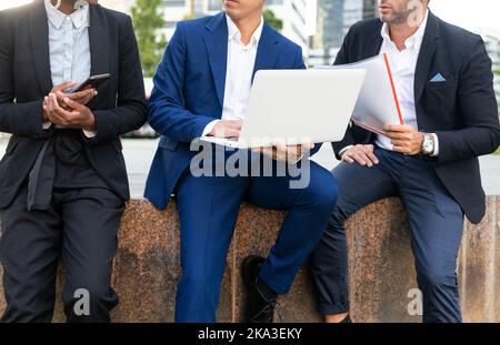 Groupe de collègues multiraciaux non reconnaissables et coupés en costumes habillés assis sur une clôture, travaillant avec un ordinateur portable et des documents tout en regardant l'écran Banque D'Images
