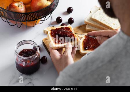 Depuis le haut de la récolte anonyme homme étalant la confiture de cerise sucrée sur des toasts croustillants avec couteau tout en préparant le petit déjeuner Banque D'Images