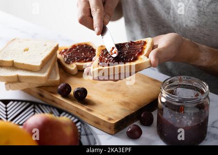 Depuis le haut de la récolte anonyme homme étalant la confiture de cerise sucrée sur des toasts croustillants avec couteau tout en préparant le petit déjeuner Banque D'Images