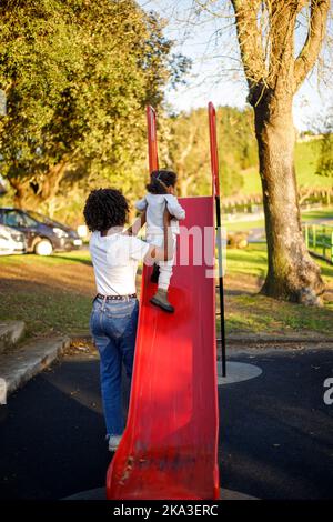 Anonyme maman noire dans des vêtements décontractés avec des cheveux bouclés soutenant la petite fille pour monter la glisse rouge le jour d'automne ensoleillé sur l'aire de jeux Banque D'Images