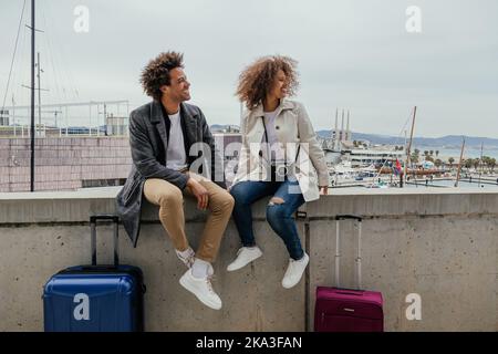 Tout le corps de touristes afro-américains, hommes et femmes, parlant tout en étant assis sur une frontière en béton près des valises dans la ville Banque D'Images