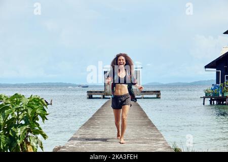 Posez une femme joyeuse en train de marcher en regardant l'appareil photo sur une jetée en bois avec des portes en profitant de la liberté sur la mer Banque D'Images