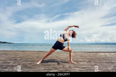 Femme hispanique à corps complet avec cheveux bouclés en casque d'activité noir exécutant Utthita Parsvakonasana sur un quai en bois de merde au soleil Banque D'Images