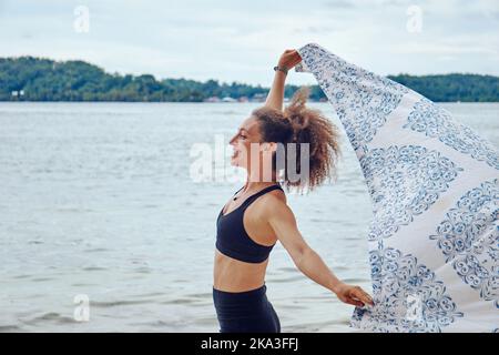 Vue latérale de la femme hispanique Happy FIT avec des cheveux bouclés tenant un linge de plage léger dans le vent debout sur la côte et regardant loin Banque D'Images