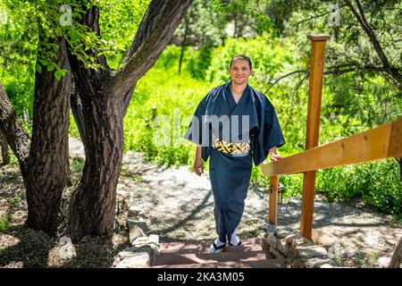 Jeune homme souriant et souriant dans un costume kimono marchant, penché sur la barrière de garde-corps dans le jardin extérieur au Japon avec vue sur la nature Banque D'Images